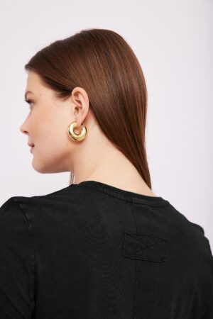 מראה צד של אישה עם שיער חום בהיר וקצר, עונדת עגילים גדולים זהובים, לובשת חולצה שחורה אופנתית.