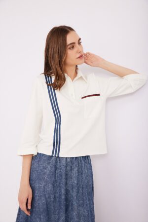 בתמונה שמלת כפתורים אופנתית לנשים בצבע לבן עם עיטור של פסים כחולים ומעין כיס בחזה, עם רקע לבן נקי.